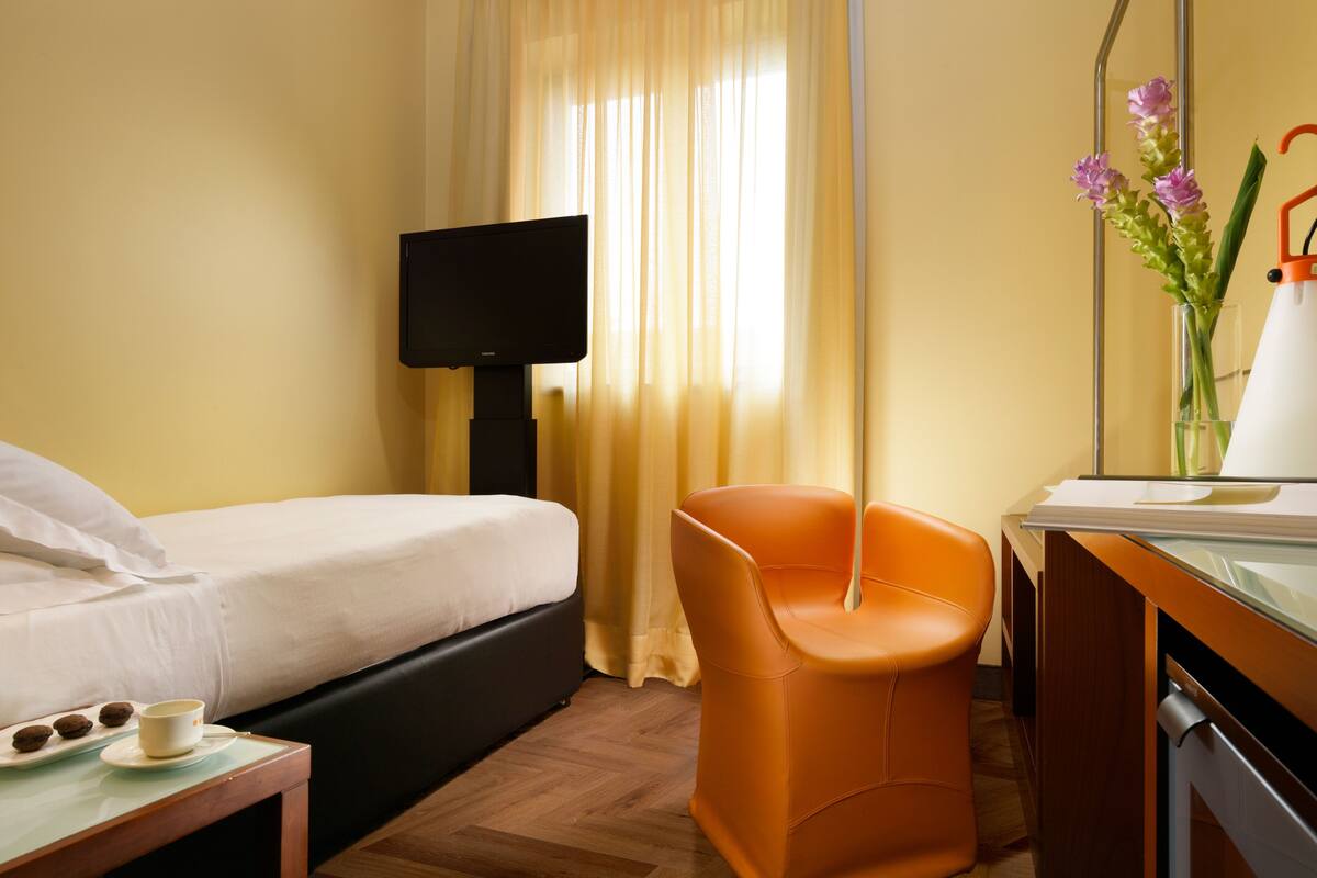 A hotel room with a bed, a desk with a chair, a coffee cup, and a vase with flowers.