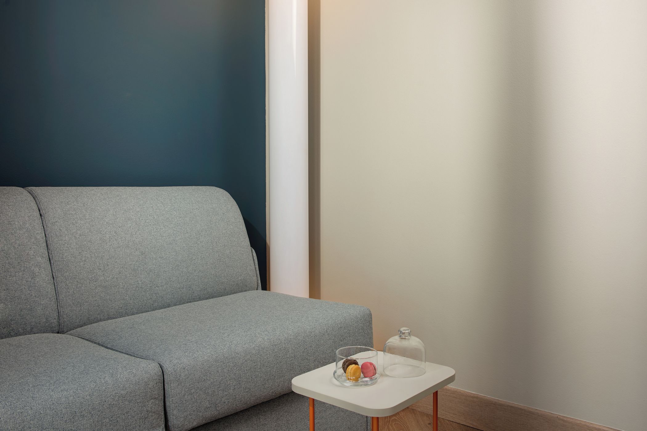 A neutral-colored sofa against a wall with a contrasting dark blue section. A small white table with a glass dome and a clear glass container with items is placed in front of the sofa.