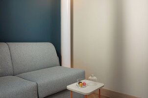 A neutral-colored sofa against a wall with a contrasting dark blue section. A small white table with a glass dome and a clear glass container with items is placed in front of the sofa.