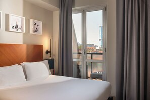 A hotel room with a large bed, a nightstand, a wall-mounted lamp, and a view of the city through a window. - UNA Hotels Mediterraneo Milano (Milan)