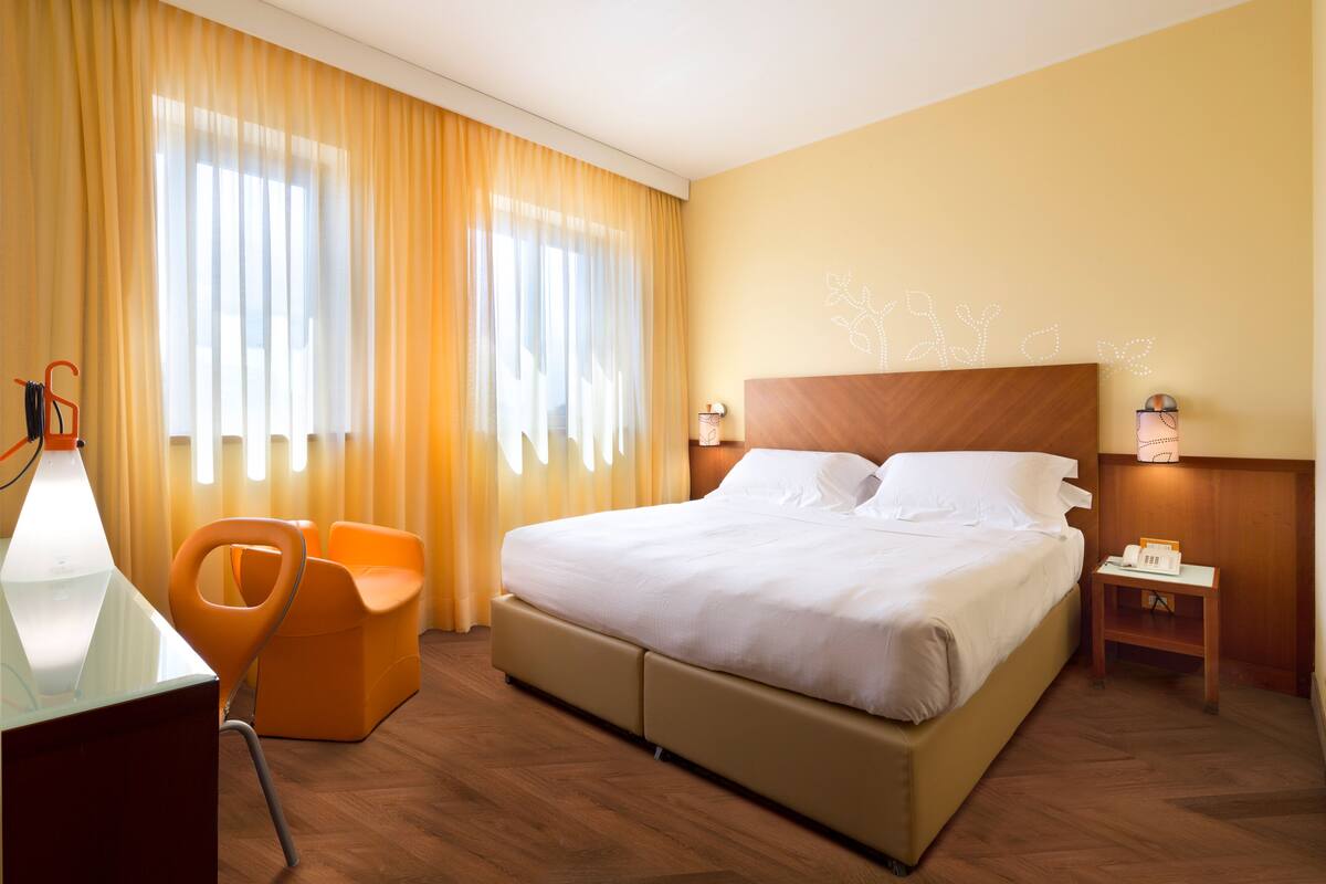 A hotel room with a large bed, a desk with a lamp, an orange chair, and a nightstand with a phone.