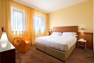 Quarto de hotel com uma cama grande, uma escrivaninha com luminária, uma poltrona laranja e uma mesa de cabeceira com telefone.