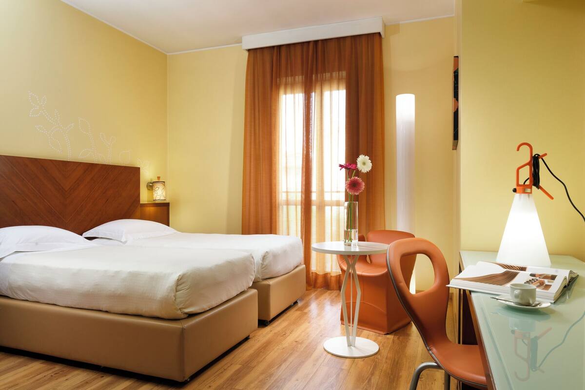A hotel room with a bed, a small table with a lamp, a chair, and a window with curtains.