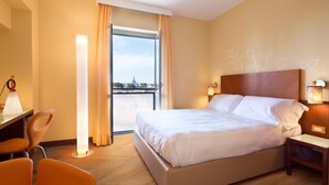 A hotel room with a bed, a desk, a chair, a nightstand, and a view of the city through a window. - UNA Hotels Mediterraneo Milano (Milan)