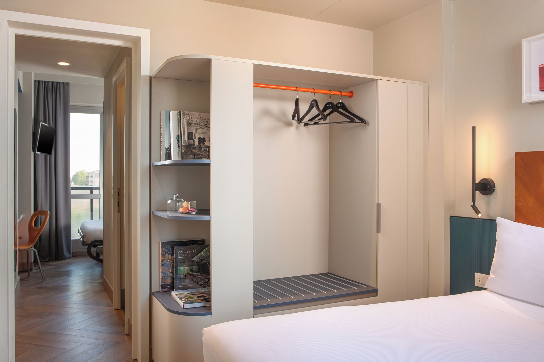 A hotel room with a bed, a seating area with a chair, a small table, a bookshelf, and a clothing rack.