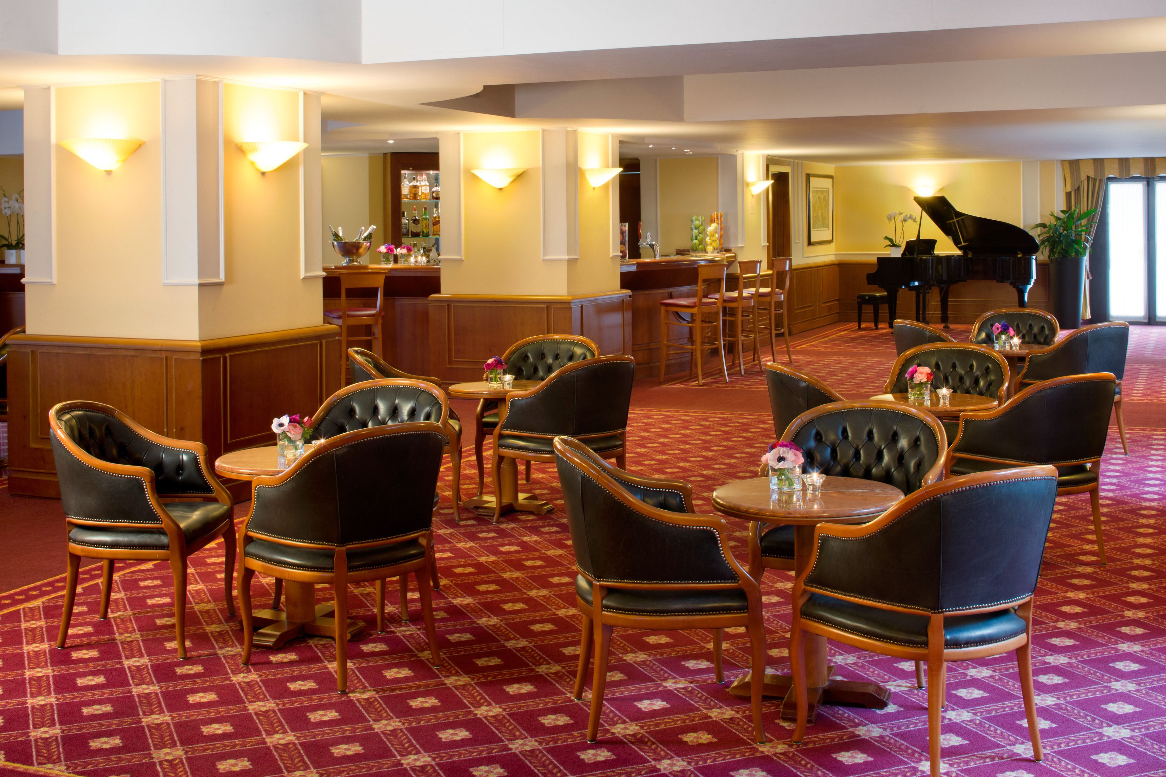 A hotel lobby with a grand piano, leather chairs, a bar area, and a patterned carpet.