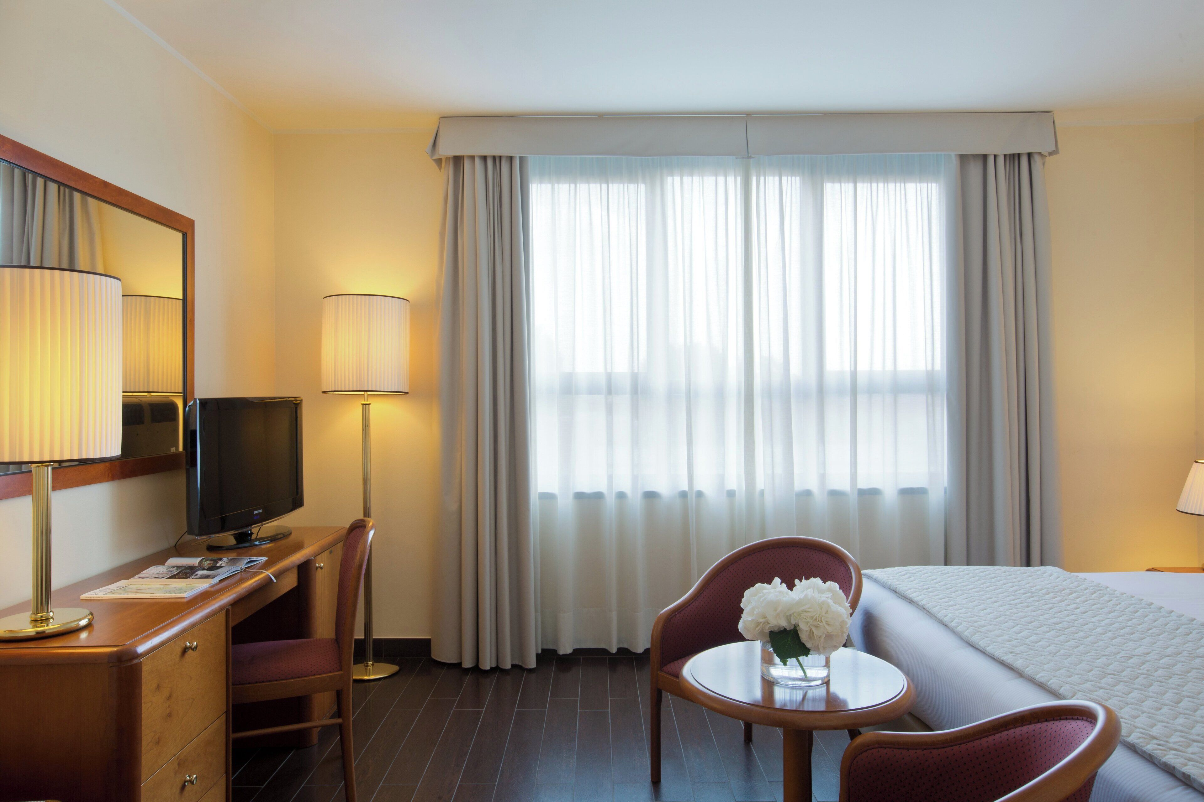 Ein Hotelzimmer mit Bett, Schreibtisch mit Fernseher, Sessel, kleinem Tisch mit Blumenvase und Fenster mit Vorhängen.