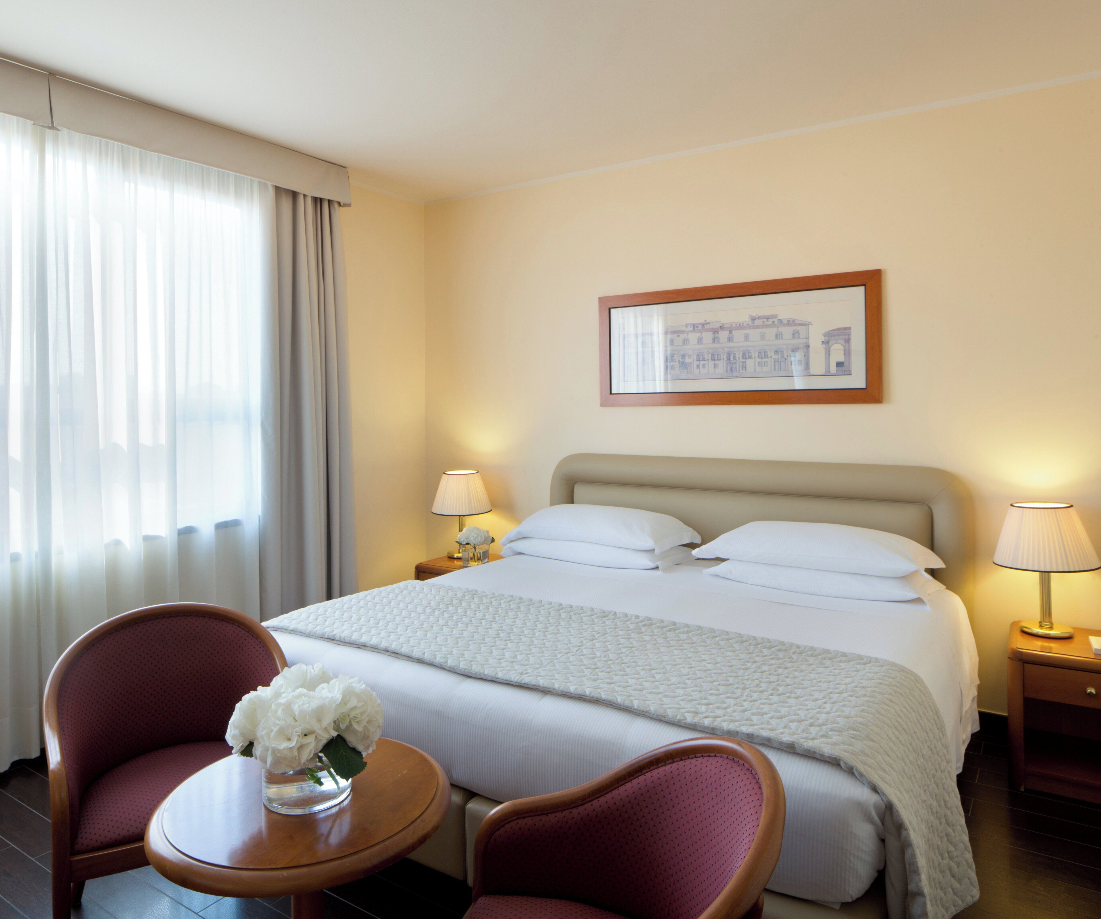 A hotel room with a bed, two bedside tables with lamps, a chair, and a window with curtains.