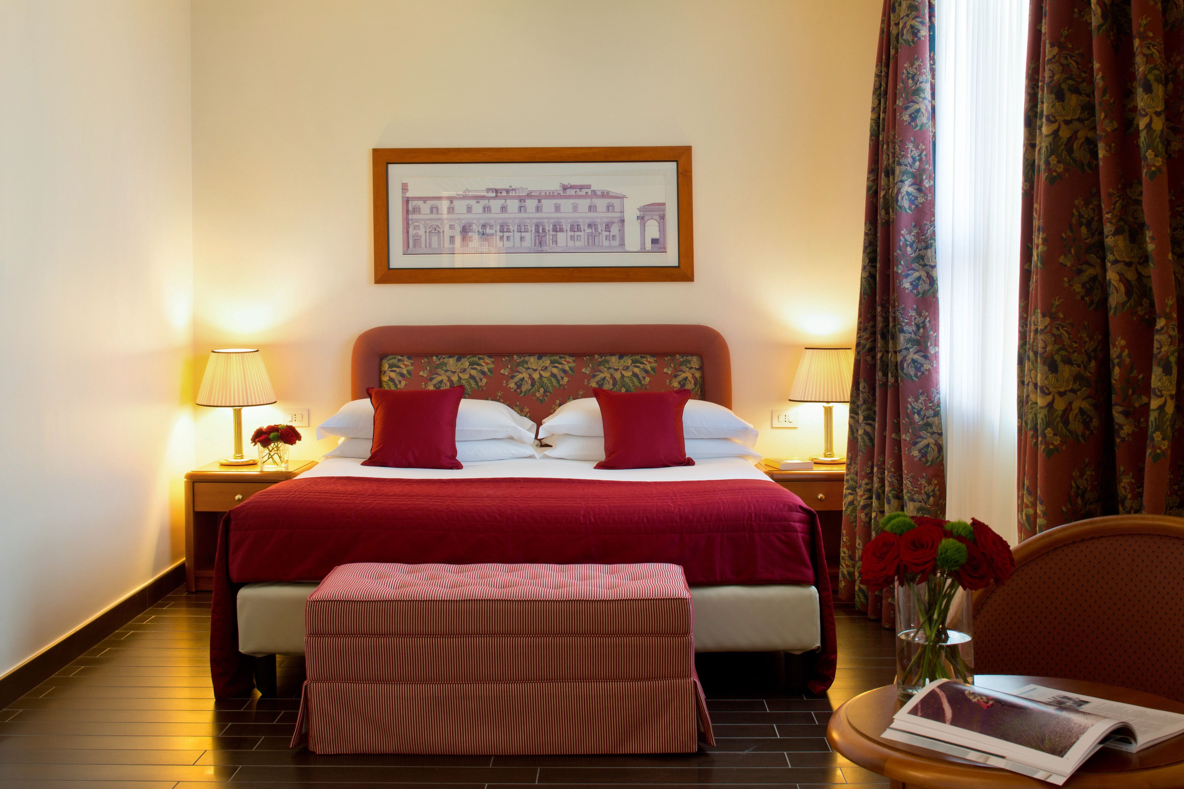 A hotel room with a large bed, two bedside lamps, a framed picture on the wall, a window with curtains, and a small table with a vase of flowers.