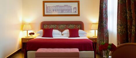 A hotel room with a large bed, two bedside lamps, a framed picture on the wall, a window with curtains, and a small table with a vase of flowers.
