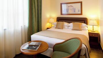 Quarto de hotel com cama, duas luminárias de cabeceira, um quadro na parede, uma cadeira e uma mesinha de centro com uma revista.