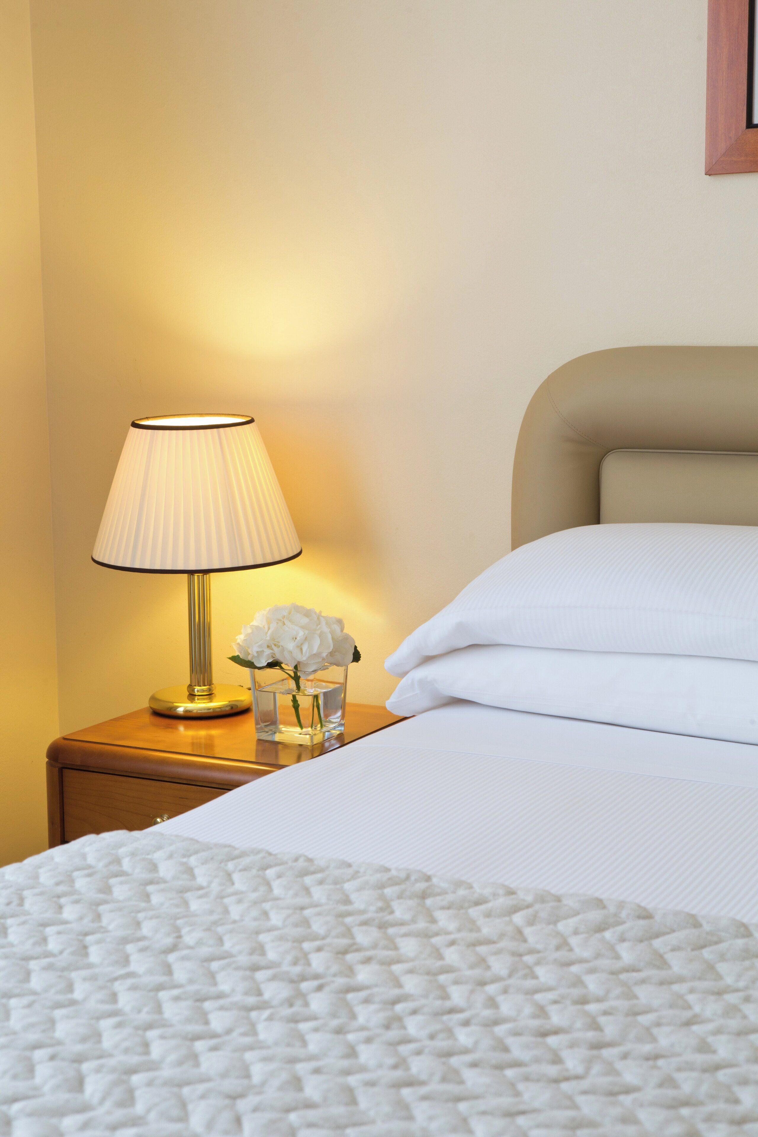 Una cama bien hecha con sábanas blancas, una lámpara de noche y un jarrón con flores blancas sobre una mesita de noche de madera.