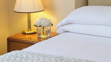 Una cama bien hecha con sábanas blancas, una lámpara de noche y un jarrón con flores blancas sobre una mesita de noche de madera.