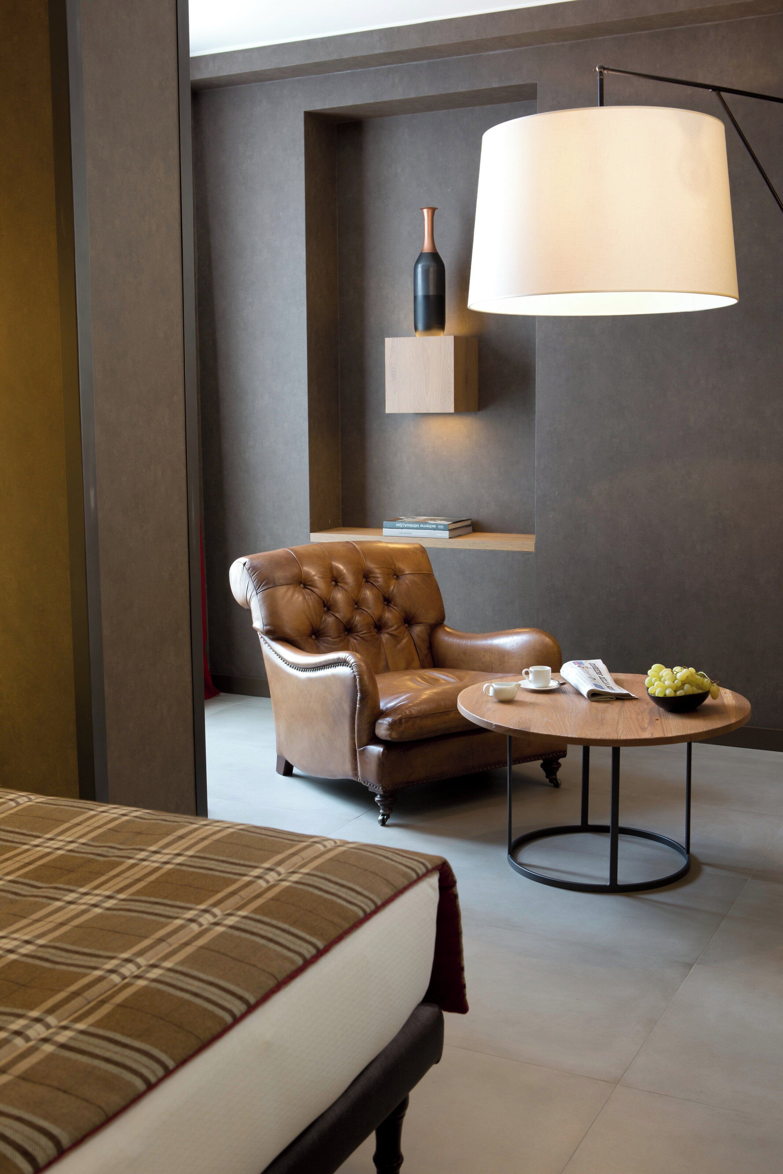 A modern hotel room with a leather armchair, a round table with a bowl of fruit, a magazine, and a bottle on a shelf.