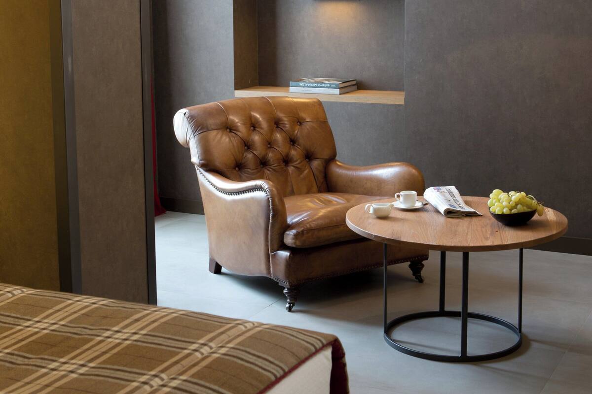 A modern hotel room with a leather armchair, a round table with a bowl of fruit, a magazine, and a bottle on a shelf.