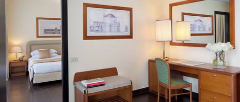 Una habitación de hotel con cama, escritorio con silla, un pequeño banco, lámpara de escritorio, espejo y cuadros en la pared.