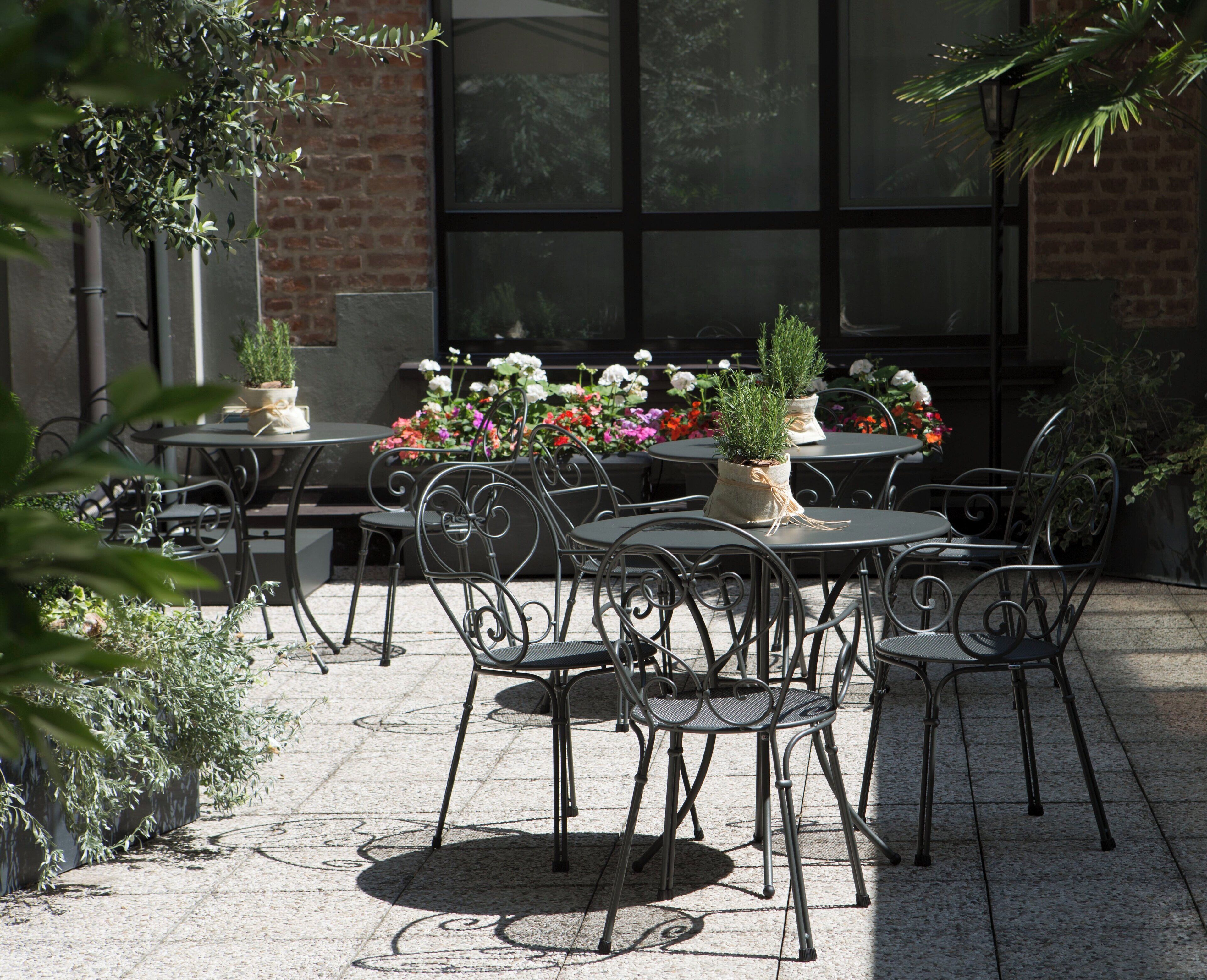 An outdoor patio with round tables and metal chairs, surrounded by potted plants and flowers.