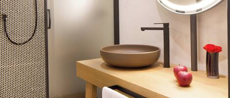 A modern bathroom with a glass-enclosed shower, a round mirror with integrated lighting, a wooden vanity, and a red apple on the counter.