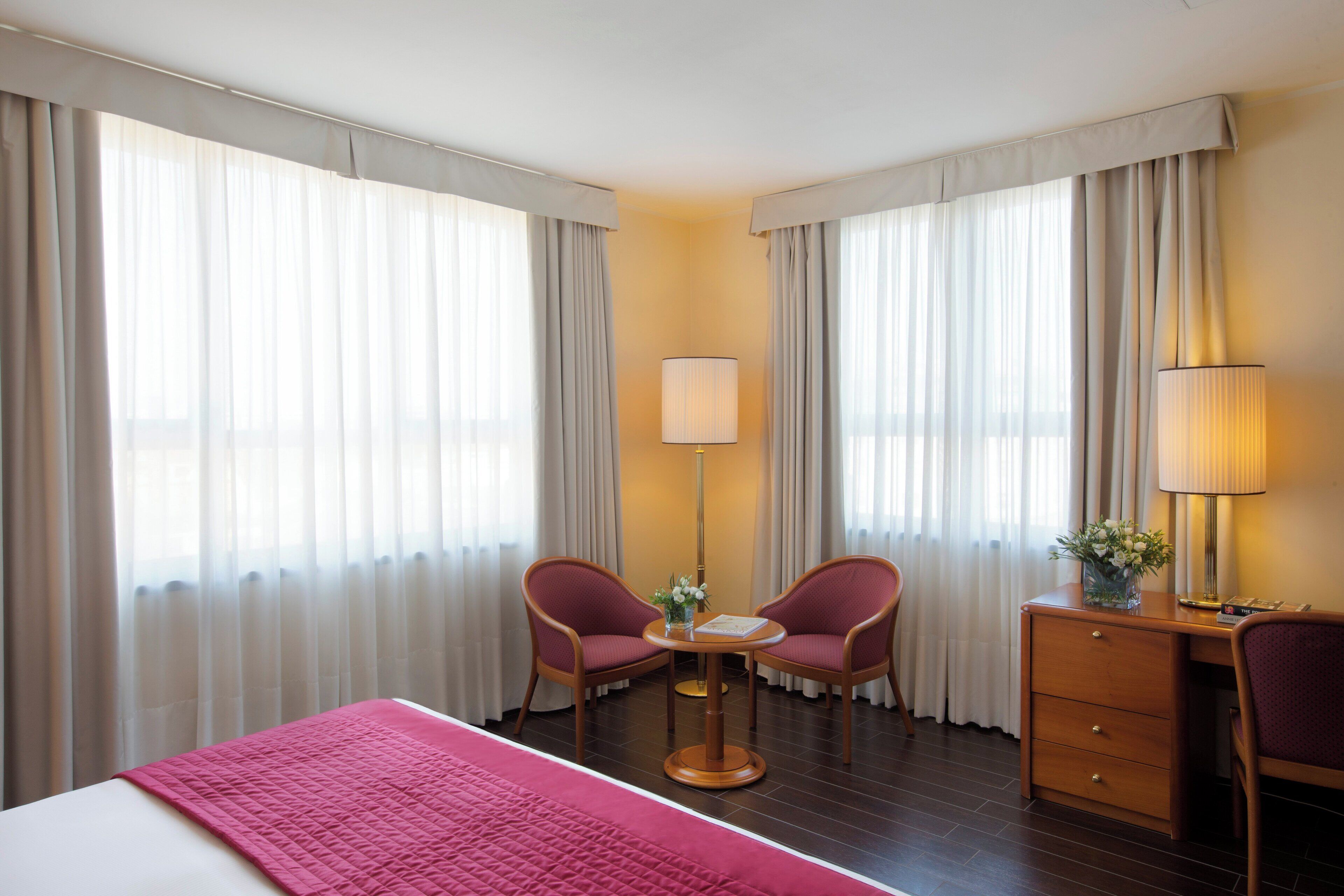 Ein Hotelzimmer mit einem Bett, zwei Sesseln, einem Schreibtisch, einer Lampe und einem Vorhangfenster.
