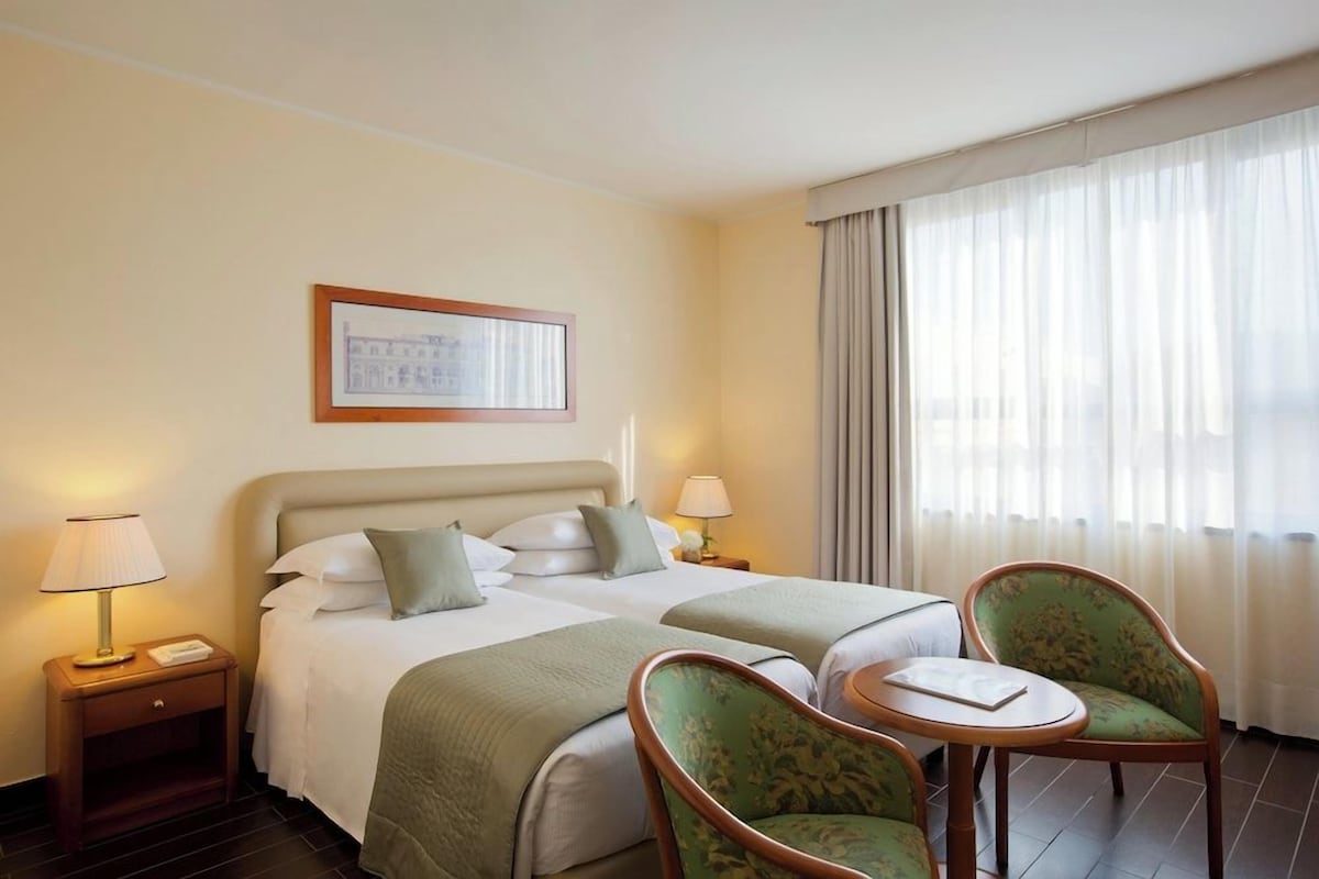 Habitación de hotel con dos camas, una mesita, dos sillas, una mesita de noche y una ventana con cortinas.