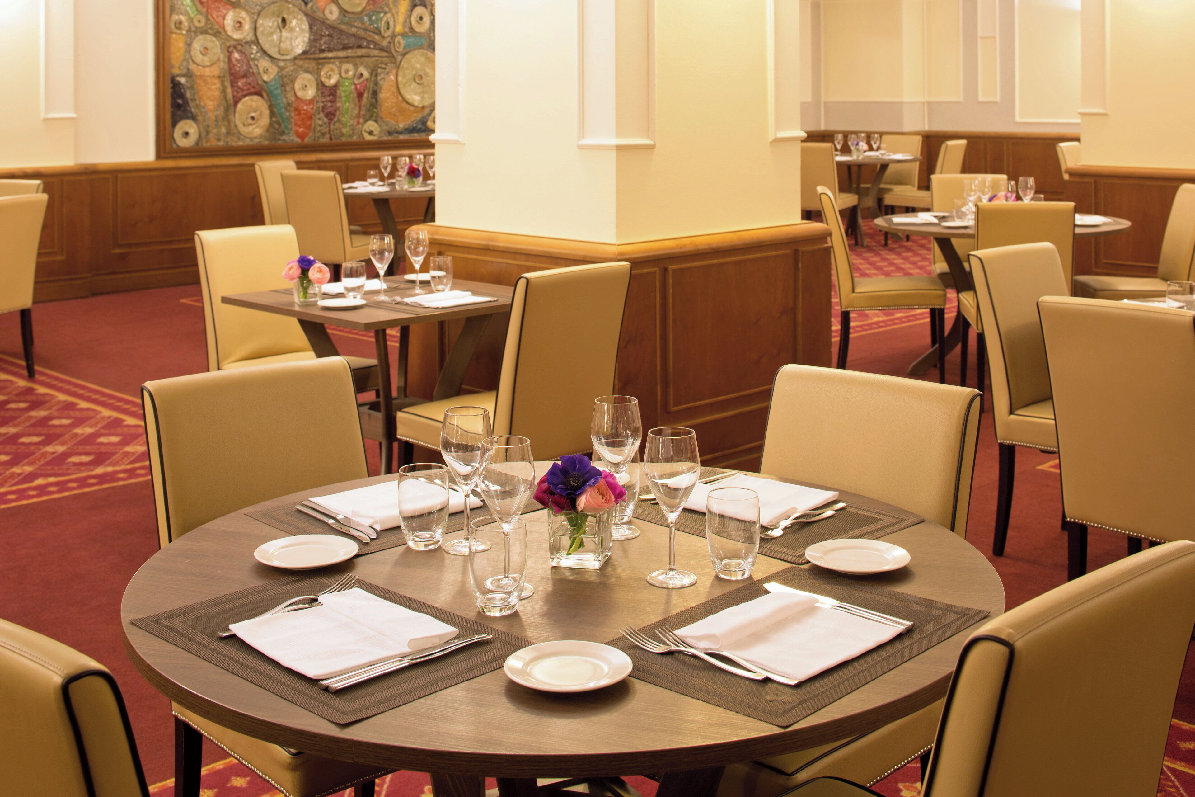 Un comedor de hotel con mesas redondas dispuestas para comer, cada una con un jarrón de flores, platos y cubiertos.