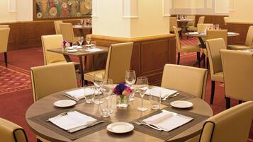 Un comedor de hotel con mesas redondas dispuestas para comer, cada una con un jarrón de flores, platos y cubiertos.