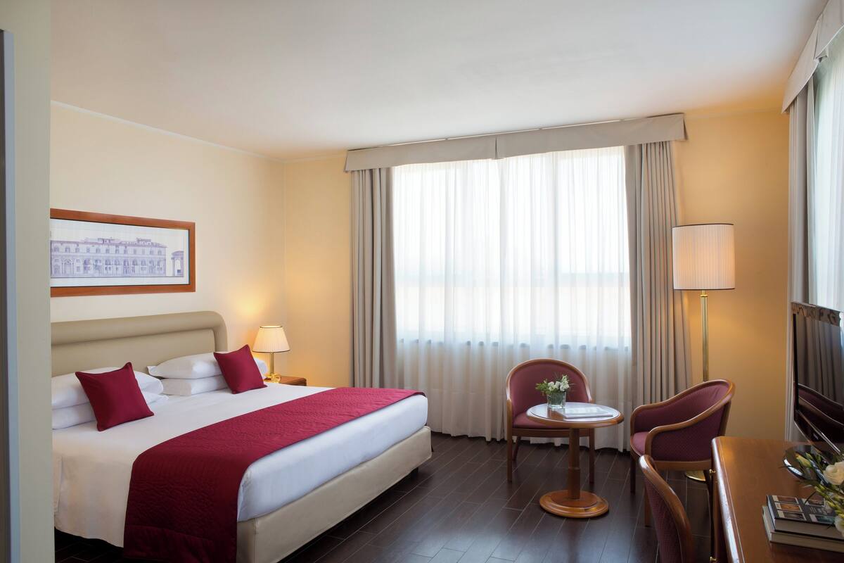 A hotel room with a large bed, a desk with a chair, a lamp, and a window with curtains.