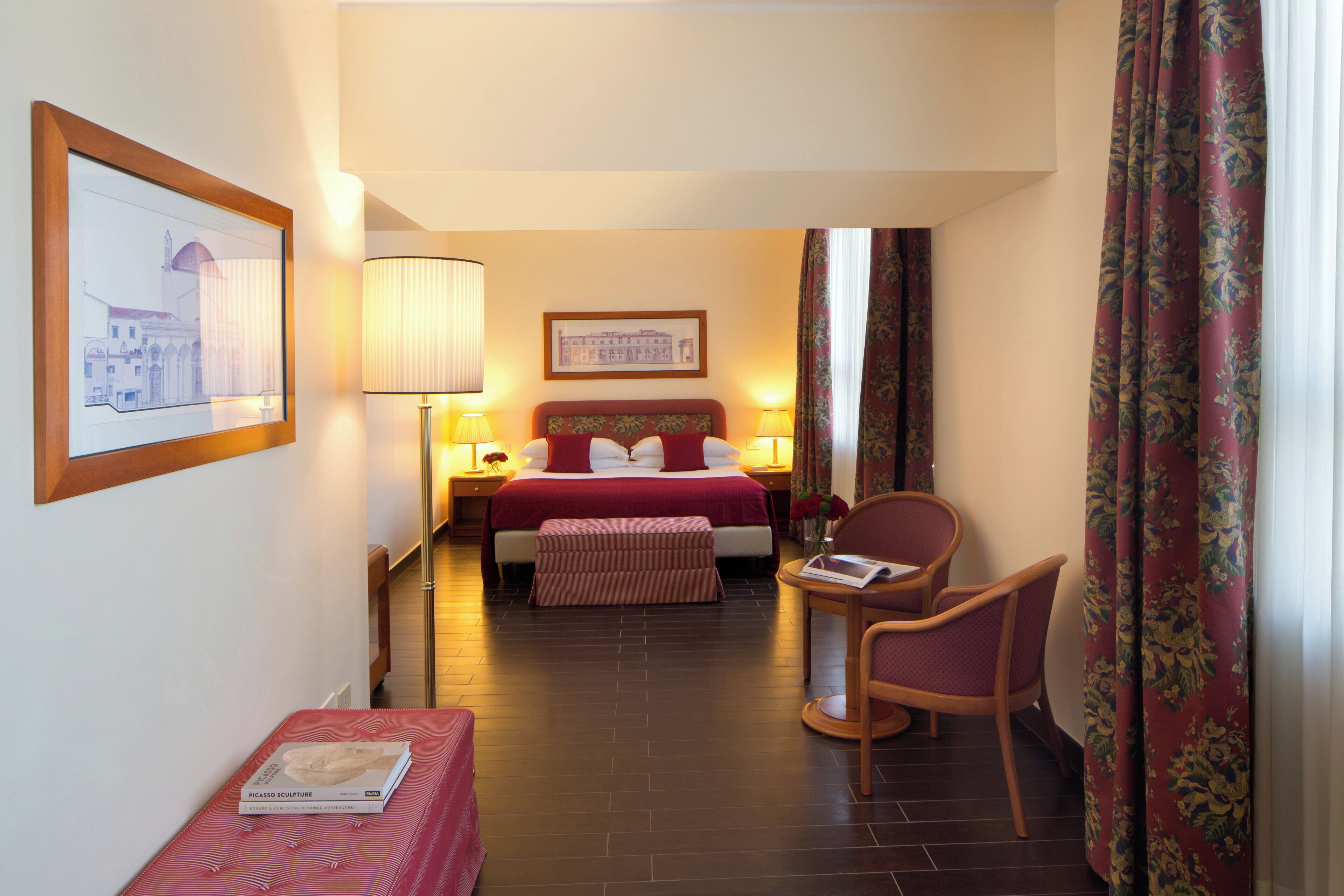 A hotel room with a bed, bedside tables, a chair, a small table, and a framed picture on the wall.