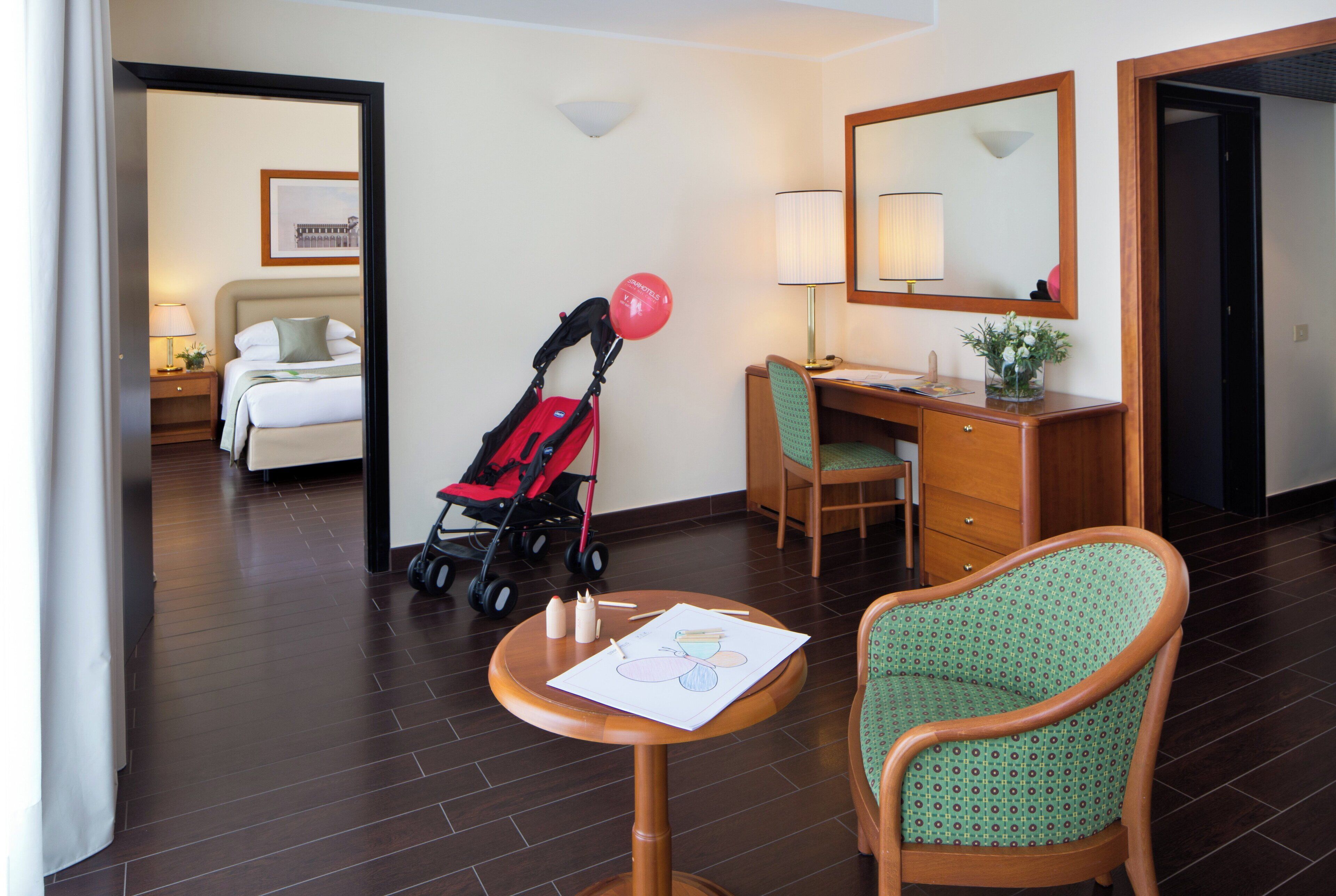 A hotel room with a bed, a desk, a chair, a table with a placemat, a lamp, a mirror, and a stroller.