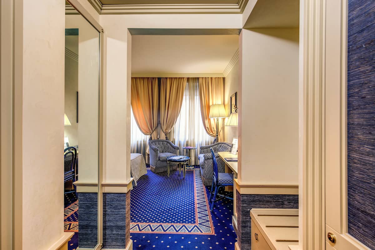 A hotel corridor with a blue patterned carpet, a seating area with chairs and a small table, and a window with curtains.