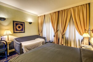 A hotel room with two beds, a desk, a chair, a lamp, and a wall-mounted artwork.