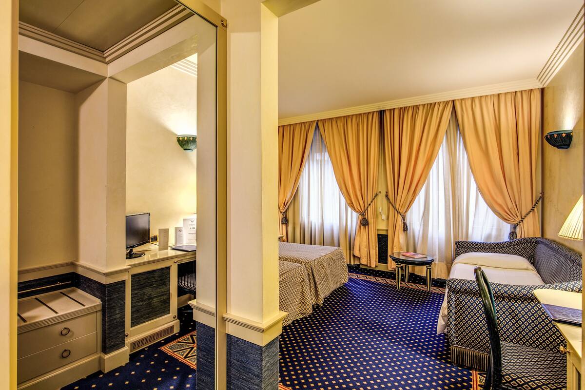 A hotel room with a bed, a desk with a computer, a chair, and a small table.