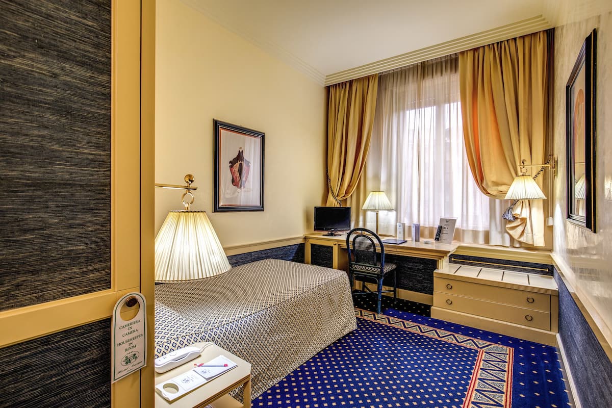 A hotel room with a bed, desk, chair, and a small table with a lamp.