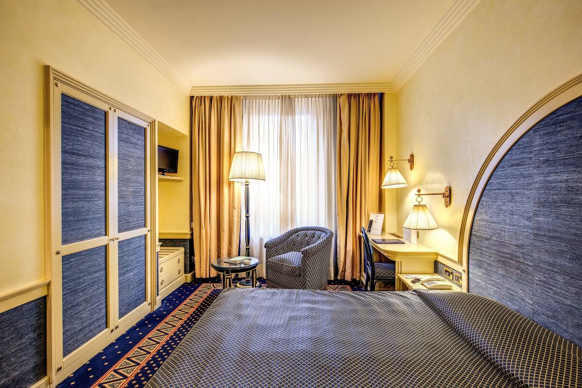 A hotel room with a large bed, a desk, a chair, a TV, and a lamp.
