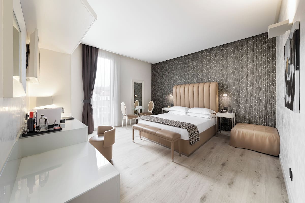 Quarto de hotel moderno com uma cama grande, uma escrivaninha, uma cadeira, um banco e uma TV fixada na parede.