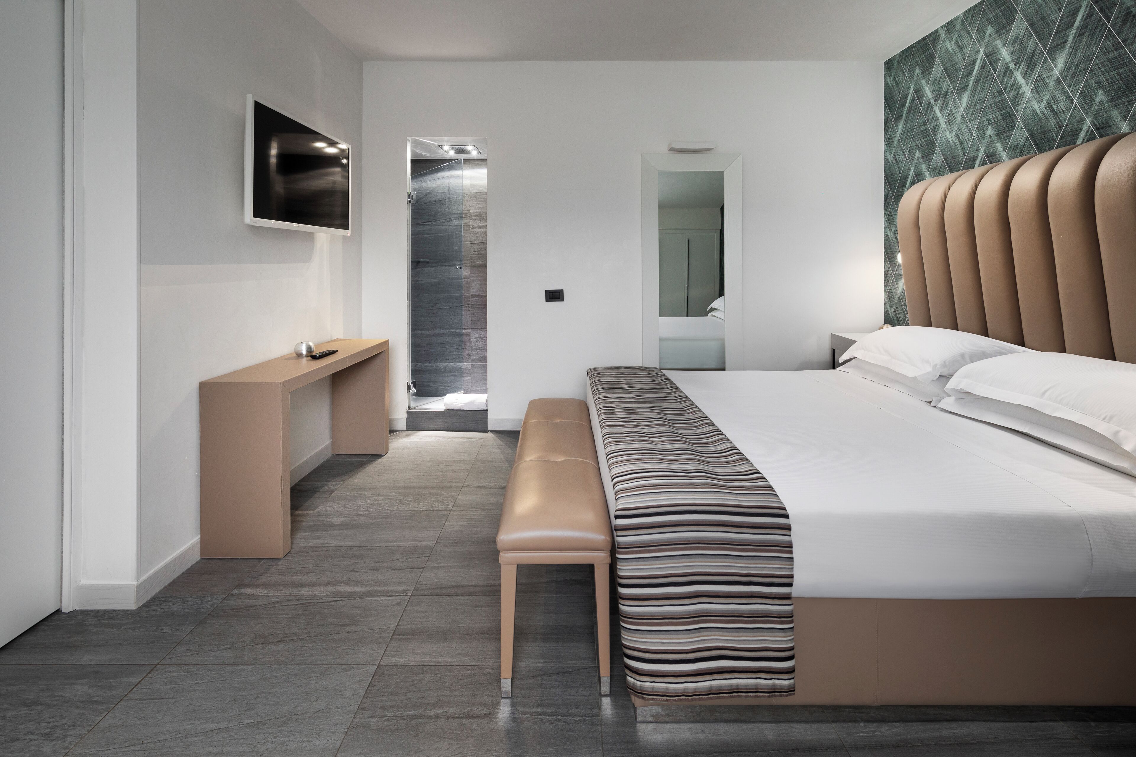 A modern bedroom with a large bed, a bench, a TV, and a bathroom visible through an open door.