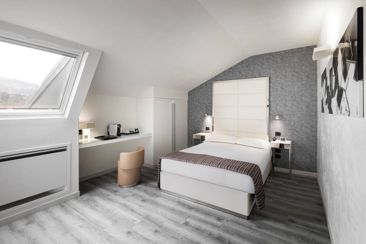 Quarto com cama, mesas de cabeceira, escrivaninha, cadeira, janela e vista para o exterior.