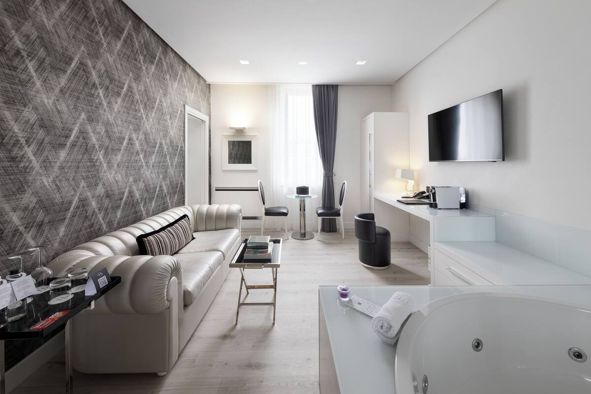 Quarto de hotel moderno com sofá, uma mesa pequena, uma mesa de centro de vidro, TV fixada na parede, banheira e uma janela grande com cortinas.