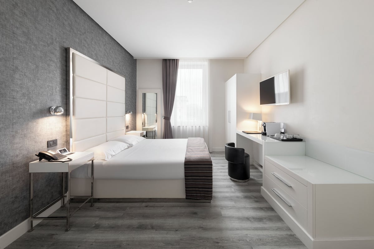 Quarto de hotel moderno com uma cama grande, mesas de cabeceira, escrivaninha, televisão e janela com cortinas.