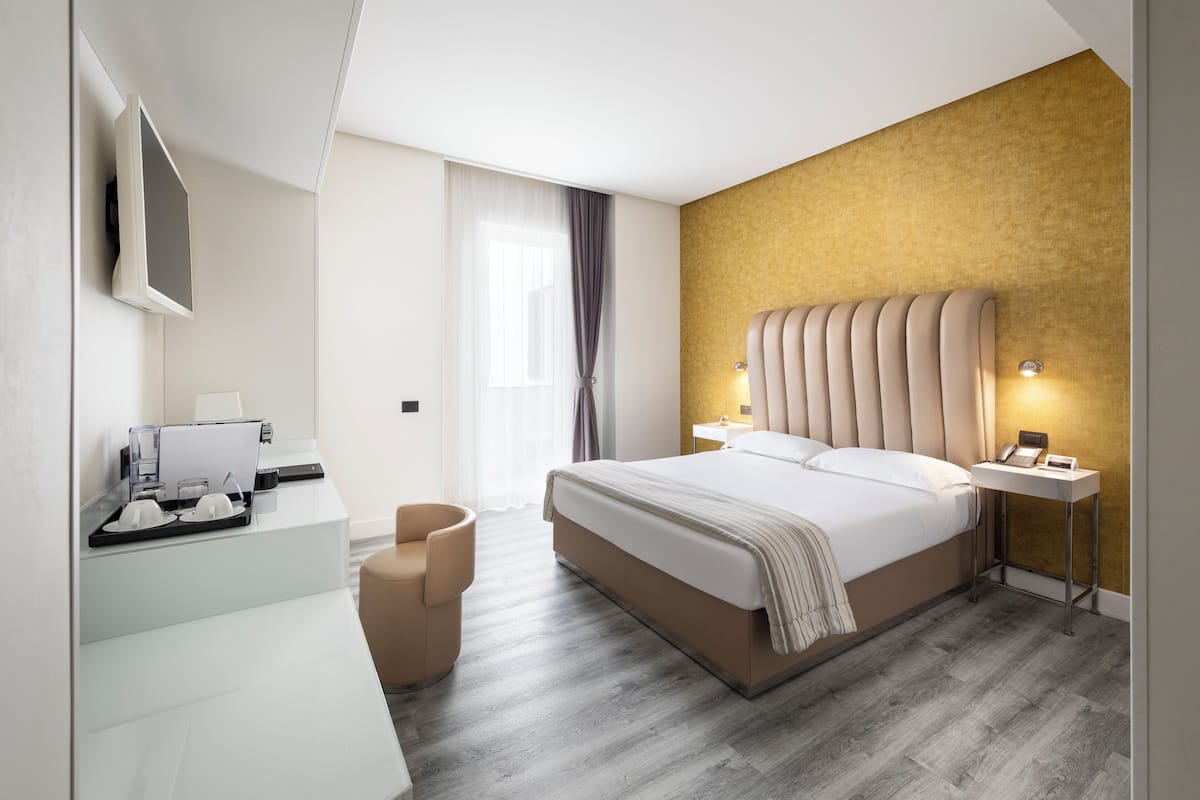 Quarto de hotel moderno com cama, mesas de cabeceira, escrivaninha com computador e televisão fixada na parede.