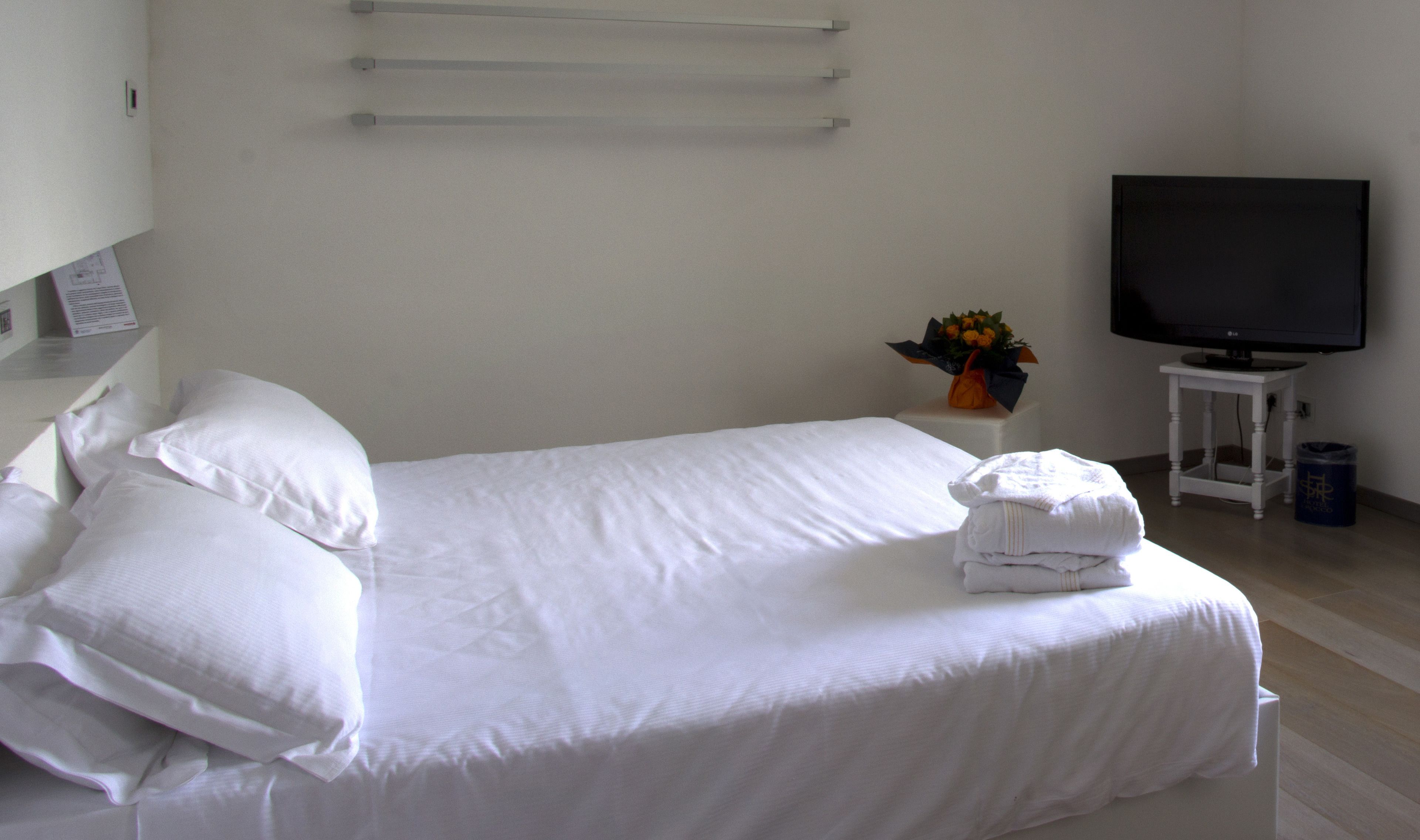 Una cama bien hecha con sábanas y almohadas blancas, una mesita pequeña con un florero de flores, y un televisor sobre un mueble.