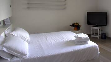 Una cama bien hecha con sábanas y almohadas blancas, una mesita pequeña con un florero de flores, y un televisor sobre un mueble.
