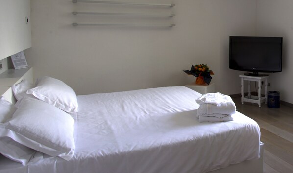 Una cama bien hecha con sábanas y almohadas blancas, una mesita pequeña con un florero de flores, y un televisor sobre un mueble.
