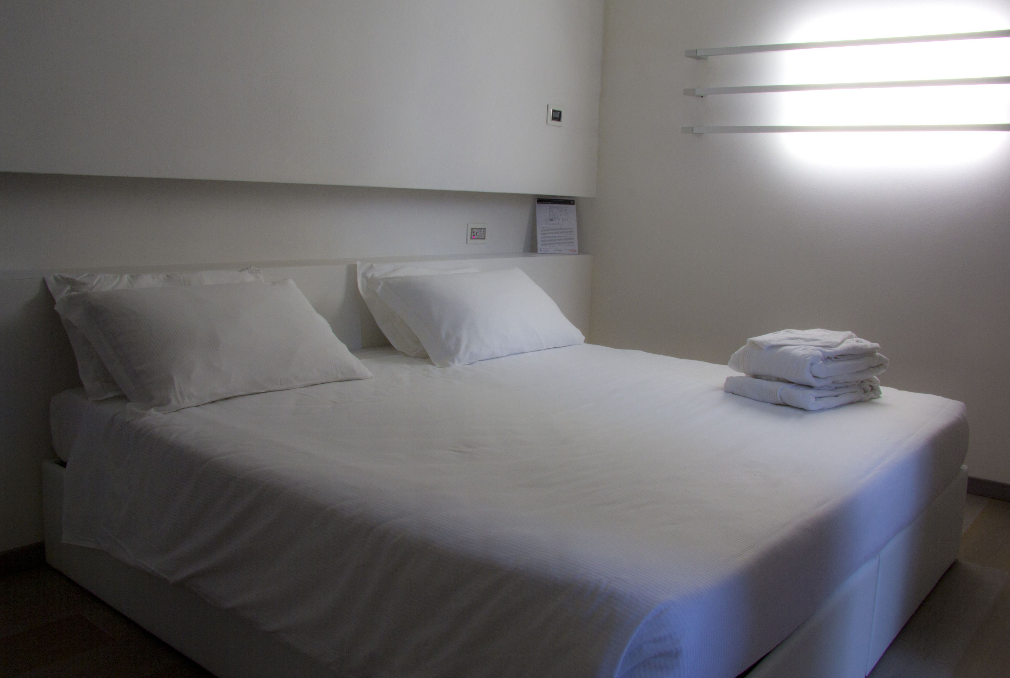 Een net opgemaakt bed met witte lakens en kussens, een stapel gevouwen handdoeken aan de zijkant, en een wandlamp.