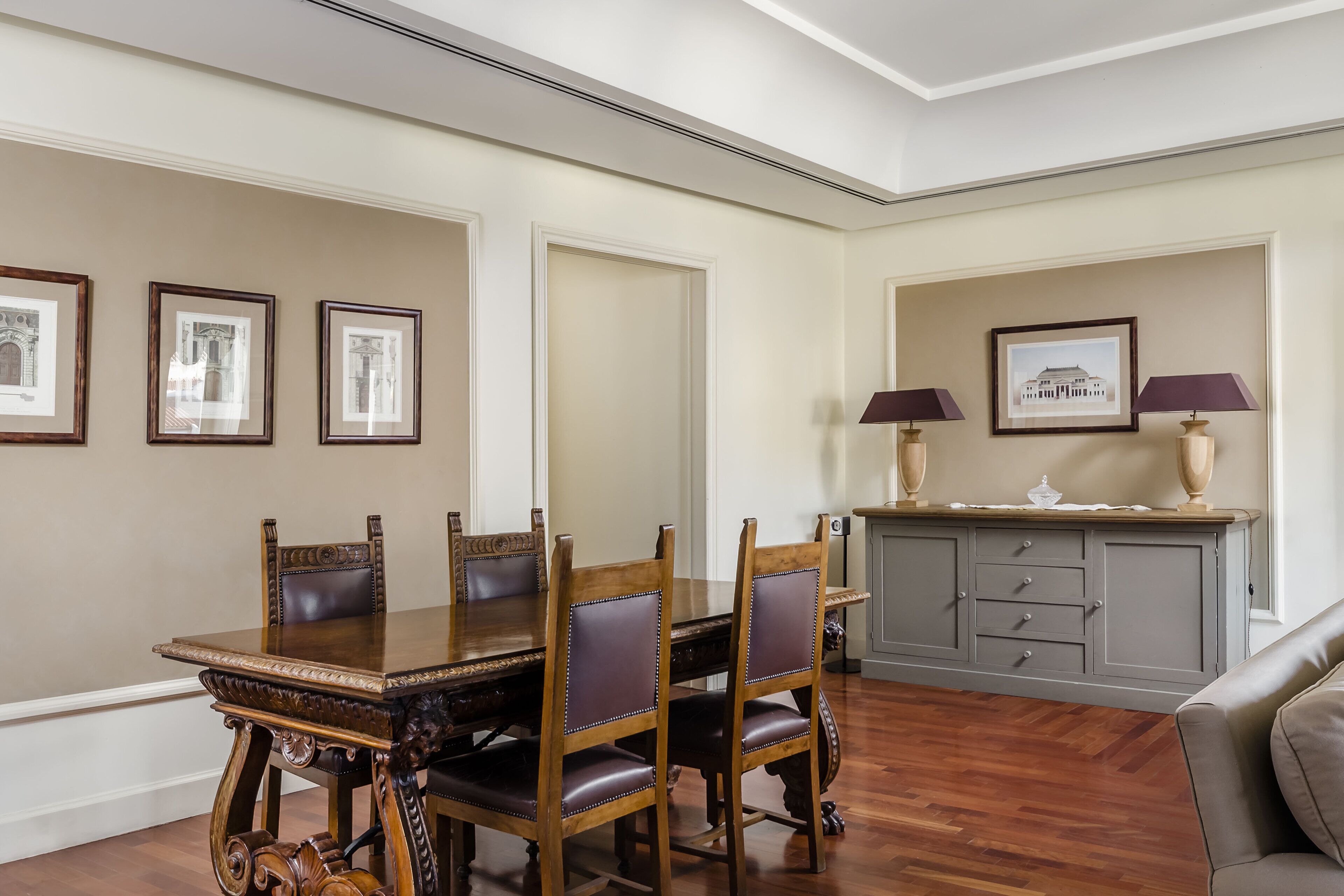 A dining room with a wooden table and chairs, framed pictures on the wall, and a grey cabinet with lamps.