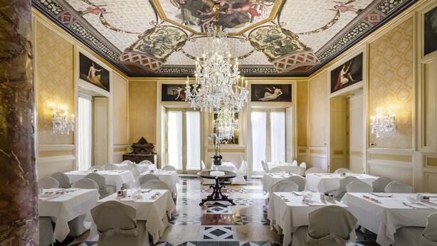 Un gran comedor con elaborada decoración en el techo, candelabros y numerosas mesas.