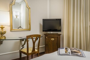 Una camera d'albergo con televisore su un mobile in legno, uno specchio a parete, una sedia e un letto con una rivista.