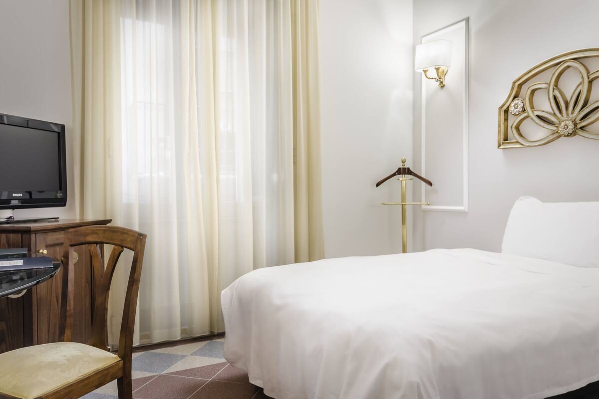A hotel room with a bed, a desk with a television, a chair, a wardrobe, and a wall-mounted light fixture.