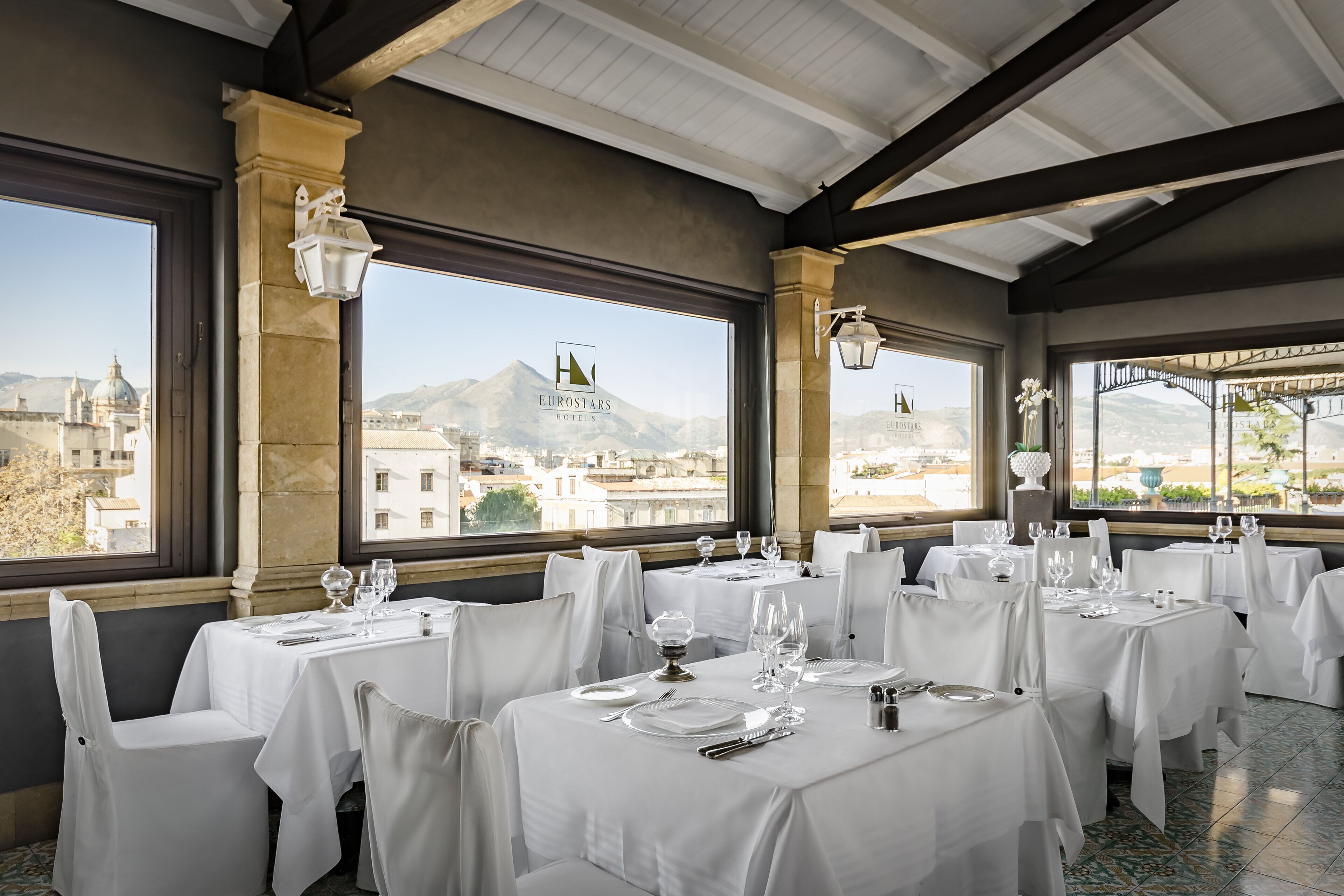 A restaurant with tables set for dining, large windows with a view of a cityscape and mountains, and elegant white tablecloths.