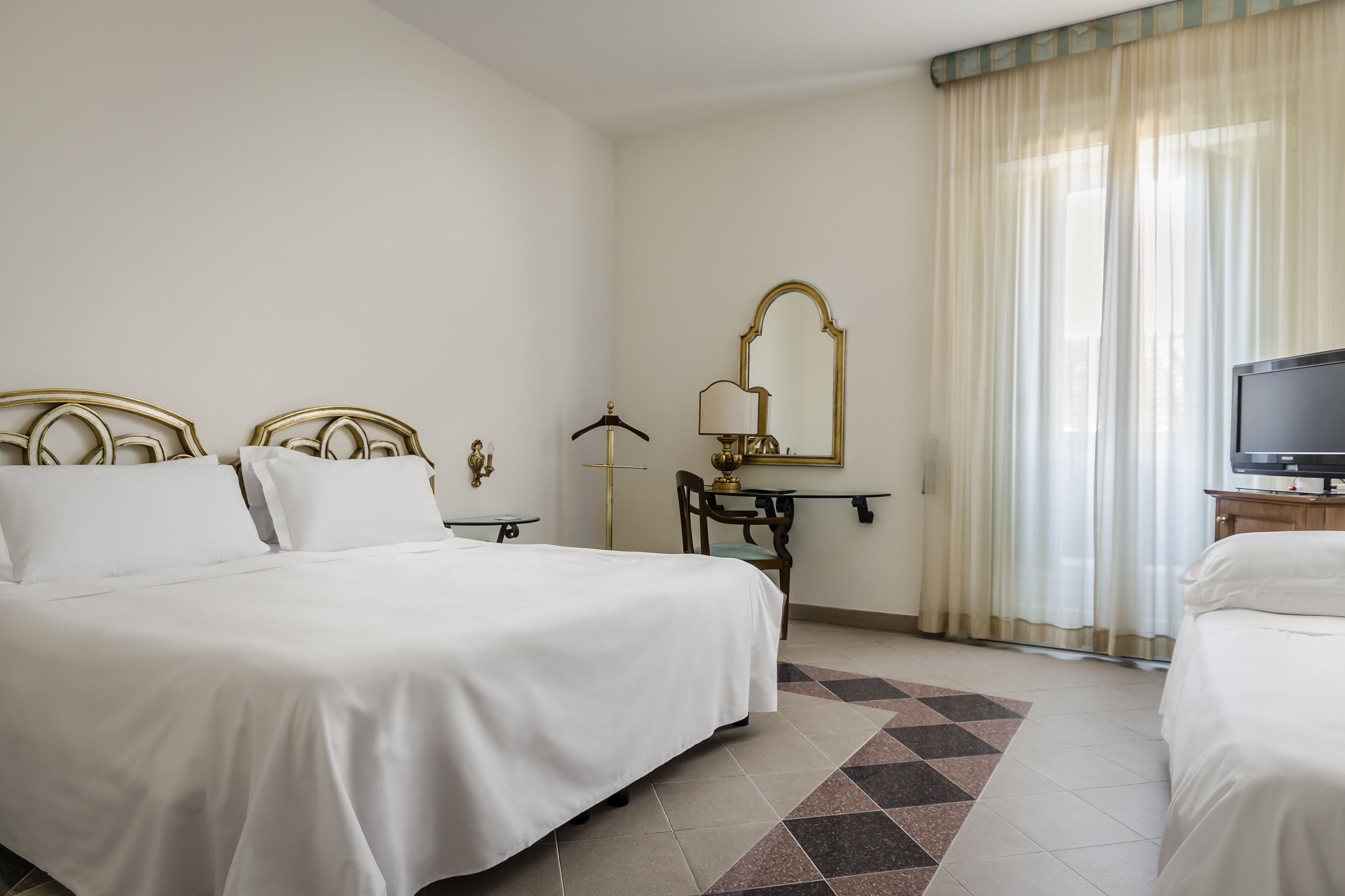 Habitación de hotel con una cama grande, una cama más pequeña, un escritorio con lámpara, un espejo y un televisor.